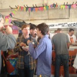 A group of partygoers eat Tijuana-style hot dogs, or TJ dogs, at the Street Food Market party March 24, 3013 in the San Diego Public Market. The banner in Spanish in the backgrounds reads, "Put your hands up and open your mouth!" (Photo by Cyndia Zwahlen for the Small Biz Mix.)